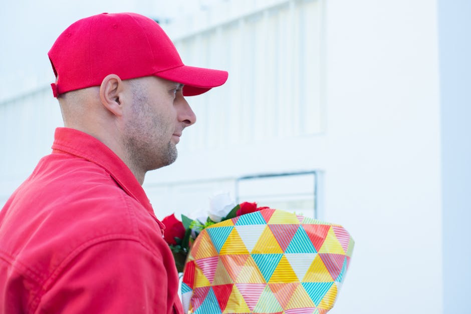 A man wearing a red cap and red jacket holds a floral gift wrapped in vibrant geometric patterned paper with yellow, blue, red, and orange triangles, accented by a red ribbon. He is standing outdoors 