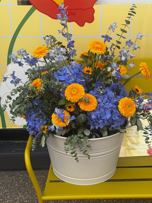 A vibrant floral arrangement featuring bright orange gerbera daisies and yellow marigolds mixed with blue hydrangeas and delphiniums, complemented by green foliage and eucalyptus stems. The bouquet is
