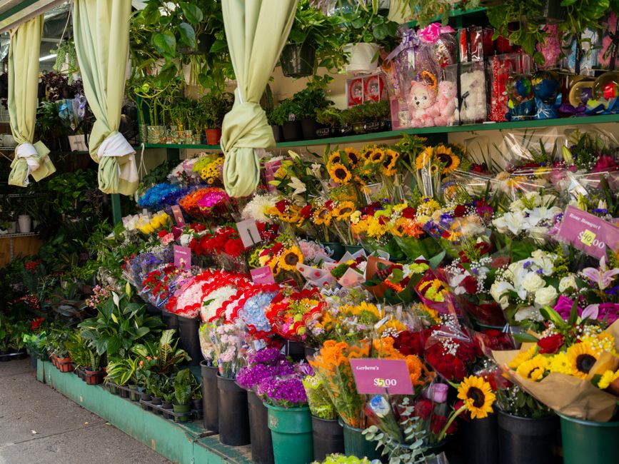 A vibrant display of fresh flower bouquets arranged on a flower stand outside a floral shop, showcasing a variety of colorful flowers including sunflowers, roses, lilies, and carnations. The bouquets 