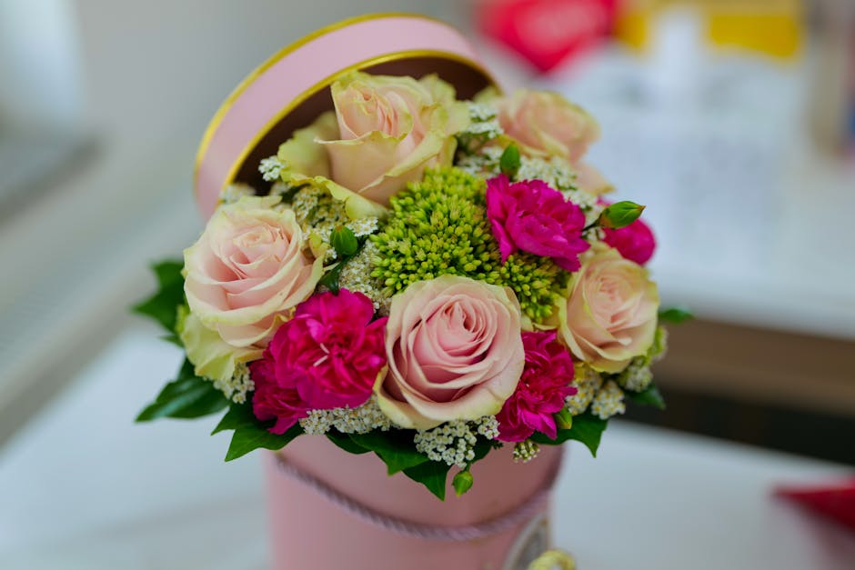 A floral arrangement featuring pale pink roses, bright magenta carnations, and small white filler flowers, all arranged in a round, pink hatbox with a subtle ribbon detail. The bouquet is rich with fr