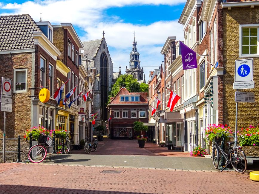 A vibrant street scene in Roehampton Lane, featuring a narrow pedestrian walkway lined with charming brick buildings, some with distinctive gabled roofs and traditional window frames. Bicycles are par