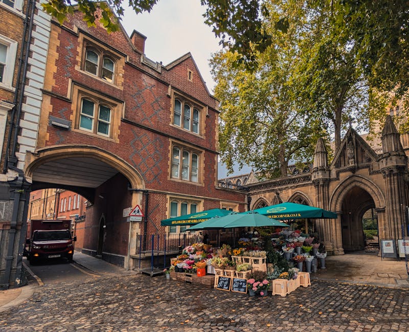 A flower stall situated on a cobblestone street beneath a large teal umbrella displaying an array of fresh floral arrangements, including pink roses, white lilies, yellow sunflowers, and assorted colo